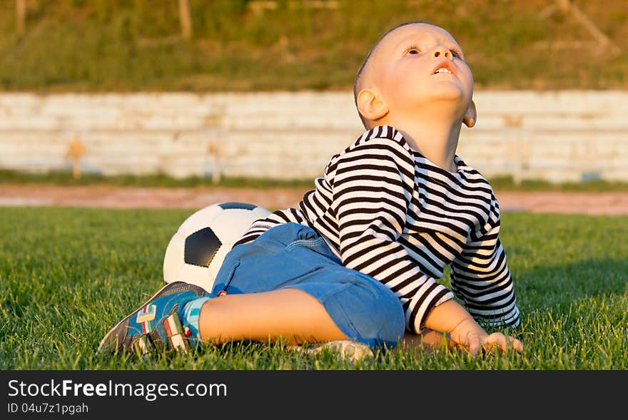 Little boy sitting on green grass with his soccer ball in evening light looking up at the sky. Little boy sitting on green grass with his soccer ball in evening light looking up at the sky