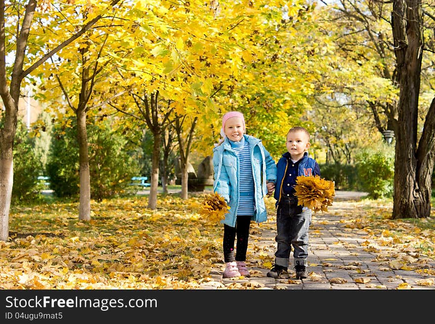 Small young brother and sister standing together on a walkway under trees in a colourful autumn park with handfuls of yellow fall leaves. Small young brother and sister standing together on a walkway under trees in a colourful autumn park with handfuls of yellow fall leaves