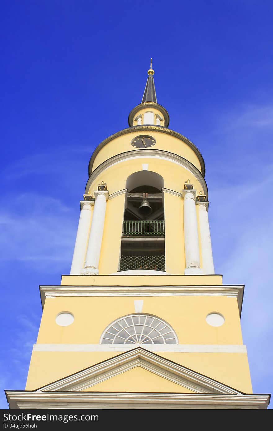 Photo of bell tower of yellow church with hours, cross, arches and columns, bells against blue morning sky. Photo of bell tower of yellow church with hours, cross, arches and columns, bells against blue morning sky