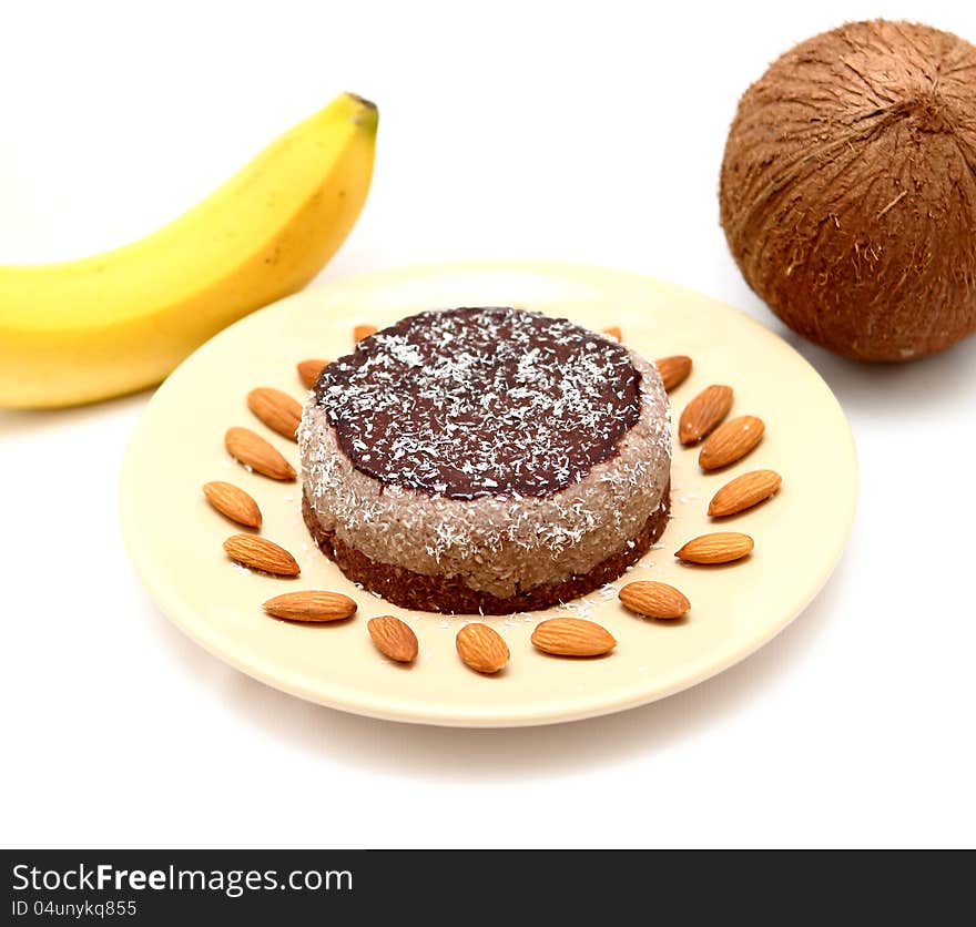 Home made cake on a brown plate covered with chocolate cream and coconut flakes on a white table with banana, almond seeds and coconut. Home made cake on a brown plate covered with chocolate cream and coconut flakes on a white table with banana, almond seeds and coconut