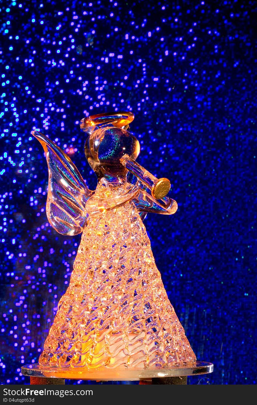 A statuette consistin of glass representing an angel which blows a trumpet. Background blue with glittering spots like stars, the glass is lit from behind with orange colored light. A statuette consistin of glass representing an angel which blows a trumpet. Background blue with glittering spots like stars, the glass is lit from behind with orange colored light