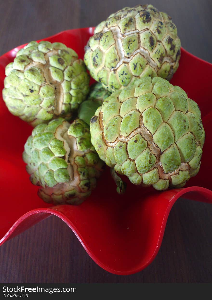 A bunch of sugar apple or Annona squamosa kept in a red bowl on a wooden table. A bunch of sugar apple or Annona squamosa kept in a red bowl on a wooden table