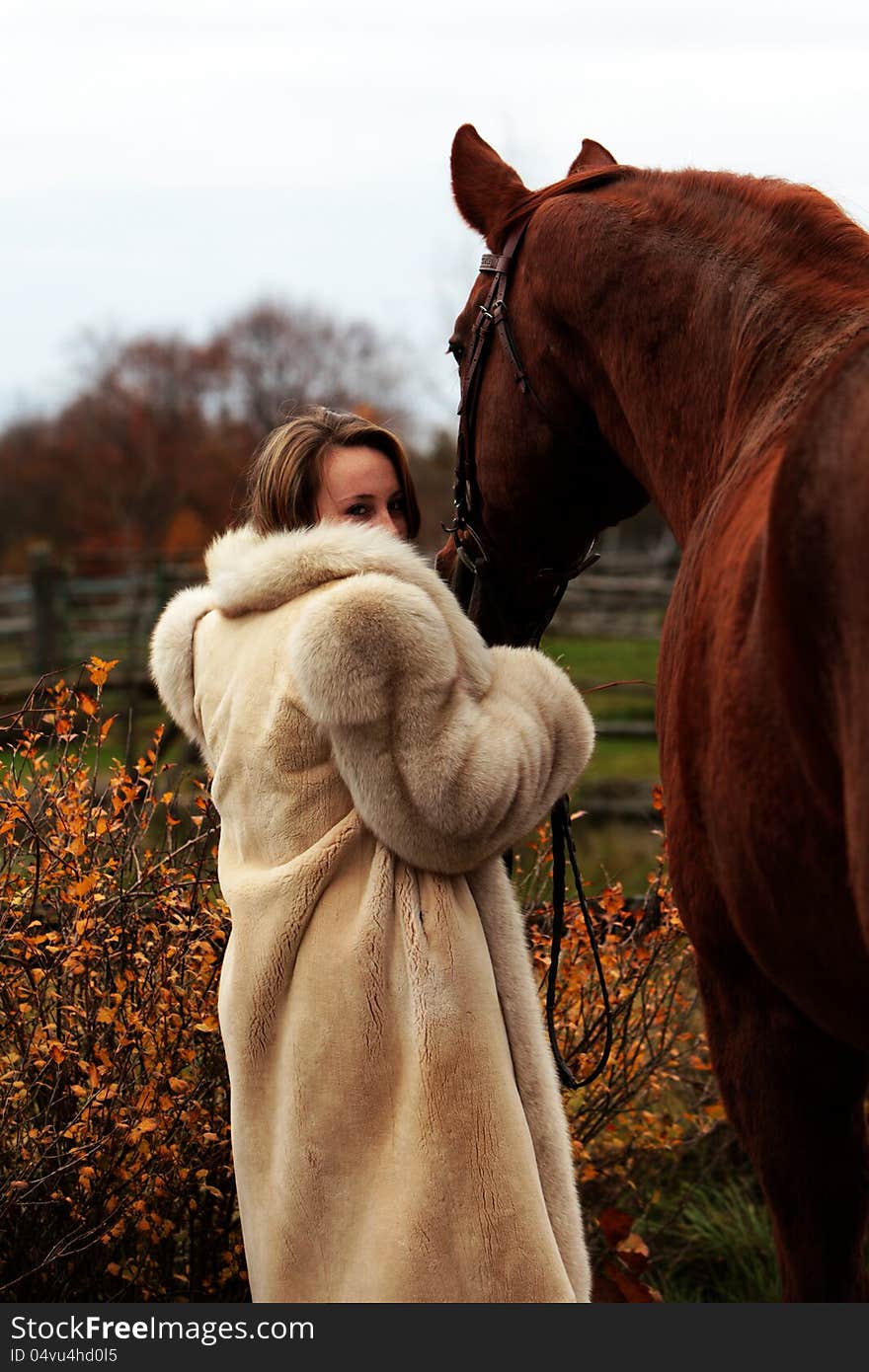 Pretty young woman in a fur coat stands holding her horse in Autumn. Pretty young woman in a fur coat stands holding her horse in Autumn.