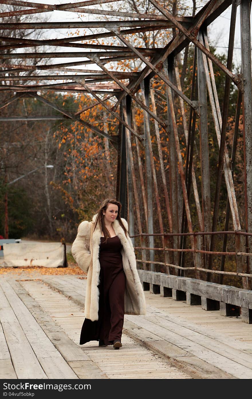 Pretty young woman wearing a long gown and fur coat goes for a walk over the old bridge in autumn. Pretty young woman wearing a long gown and fur coat goes for a walk over the old bridge in autumn.