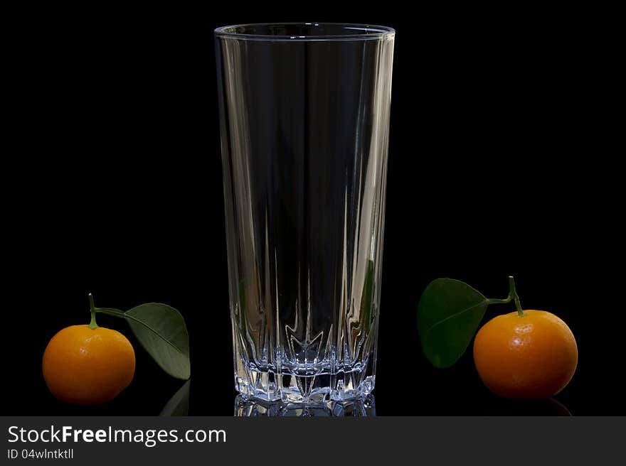 The composition of the three objects on a black background, one empty glass of clear glass and the right and left of the cup bright orange tangerines. Each mandarin on petiole have a green leaf. The composition of the three objects on a black background, one empty glass of clear glass and the right and left of the cup bright orange tangerines. Each mandarin on petiole have a green leaf.