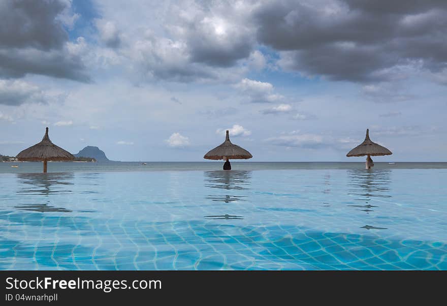 Low angle landscape image of a bright blue infinity pool against a stormy cloudy sky framed by 3 thatched beach umbrellas.