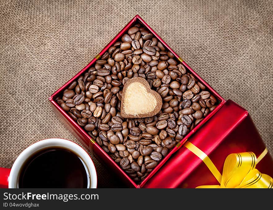 Coffee grains with heart shape cookie in Red box with yellow ribbon and mug with black coffee on textured brown background on Valentines day. Coffee grains with heart shape cookie in Red box with yellow ribbon and mug with black coffee on textured brown background on Valentines day