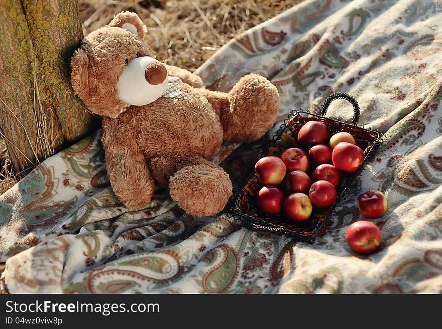 Under a tree on a blanket sitting bear with red apples. Under a tree on a blanket sitting bear with red apples