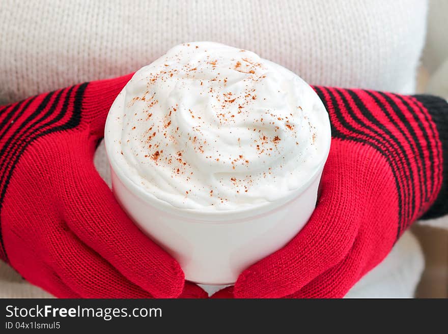 Seasonal drink topped with whipped cream and cinnamon held by person wearing red wool gloves. Seasonal drink topped with whipped cream and cinnamon held by person wearing red wool gloves