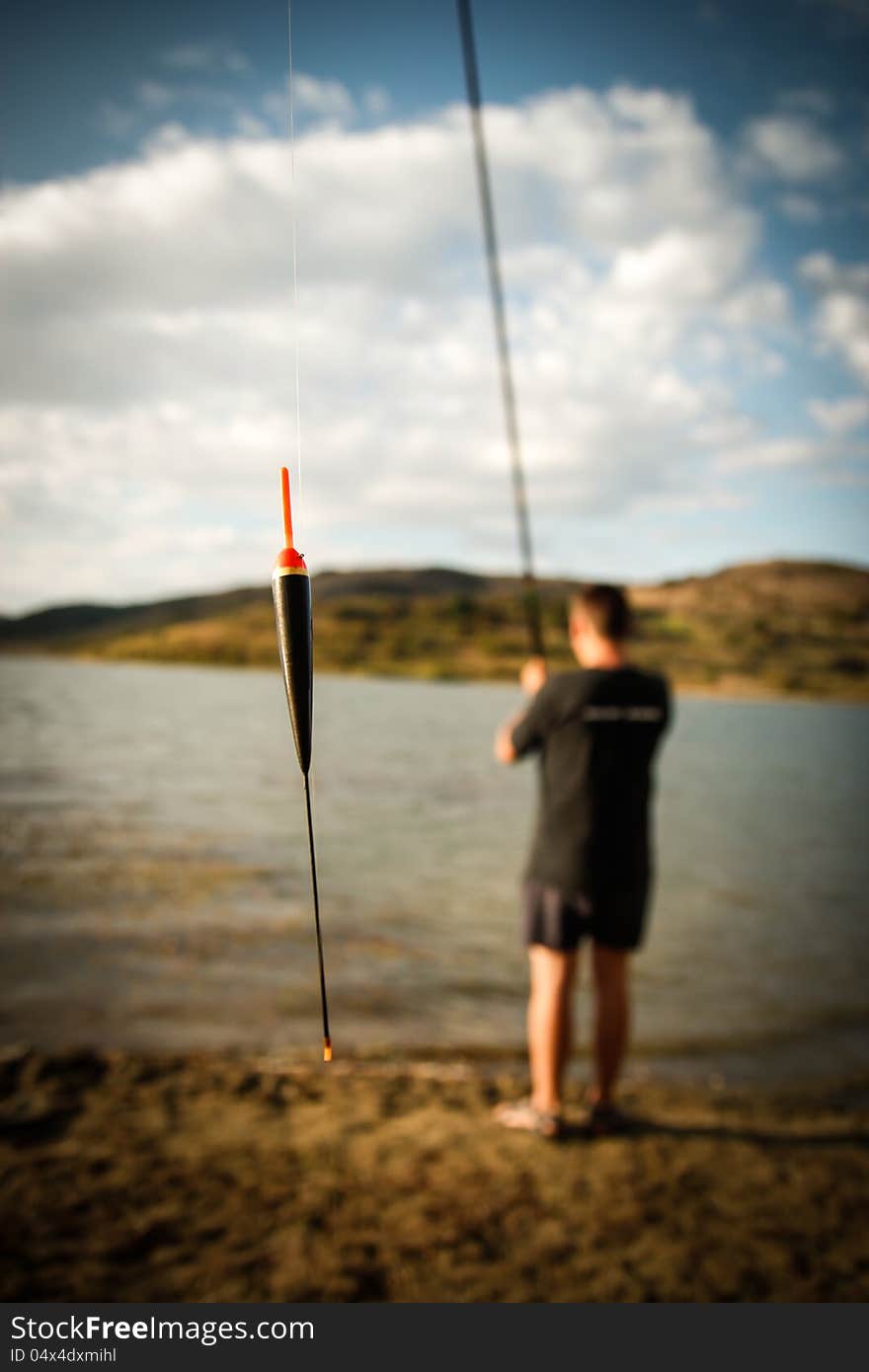 Lakeside fishing, focus on floater hanging from the line, the background is out of focus having the fisherman standing with the fishing rod facing away toward the lake