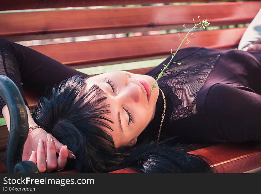 Young woman enjoying the sun, lying on a bench, with closed eyes and a flower in her lips. Young woman enjoying the sun, lying on a bench, with closed eyes and a flower in her lips