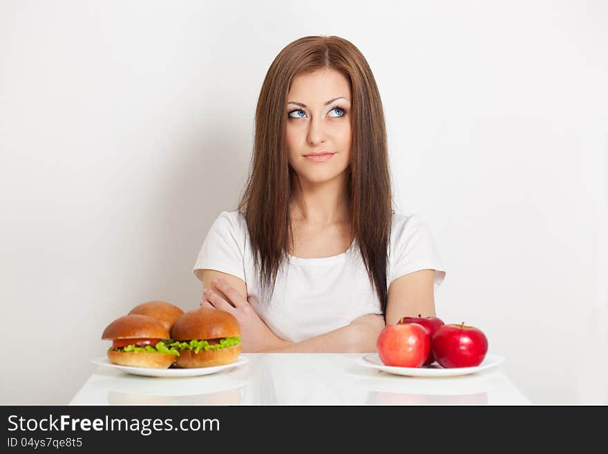 Woman sitting behind the table with standing junk food and apples and thinking what to choose. Woman sitting behind the table with standing junk food and apples and thinking what to choose