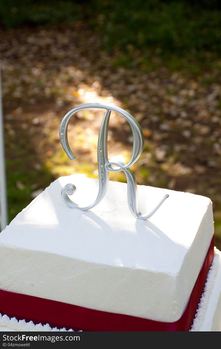 A white and red square wedding cake with multiple tiers and the letter R on top sits outside on a yellow table for a wedding reception. A white and red square wedding cake with multiple tiers and the letter R on top sits outside on a yellow table for a wedding reception.