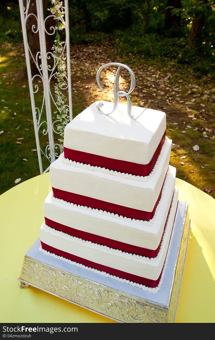 A white and red square wedding cake with multiple tiers and the letter R on top sits outside on a yellow table for a wedding reception. A white and red square wedding cake with multiple tiers and the letter R on top sits outside on a yellow table for a wedding reception.