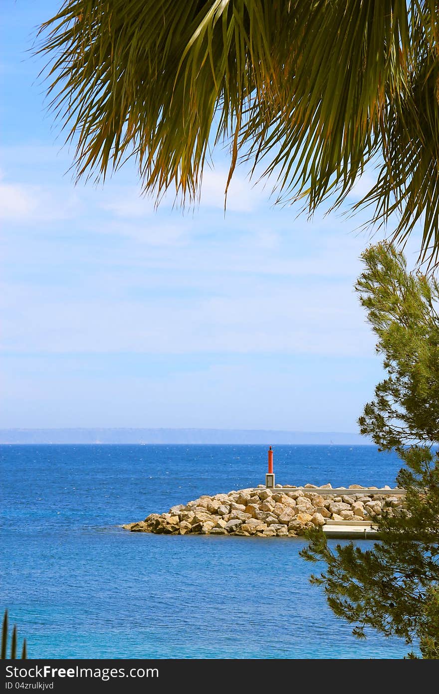 Sea view through a frame formed by palm tree and other plants, rock pier with red sign pole visible