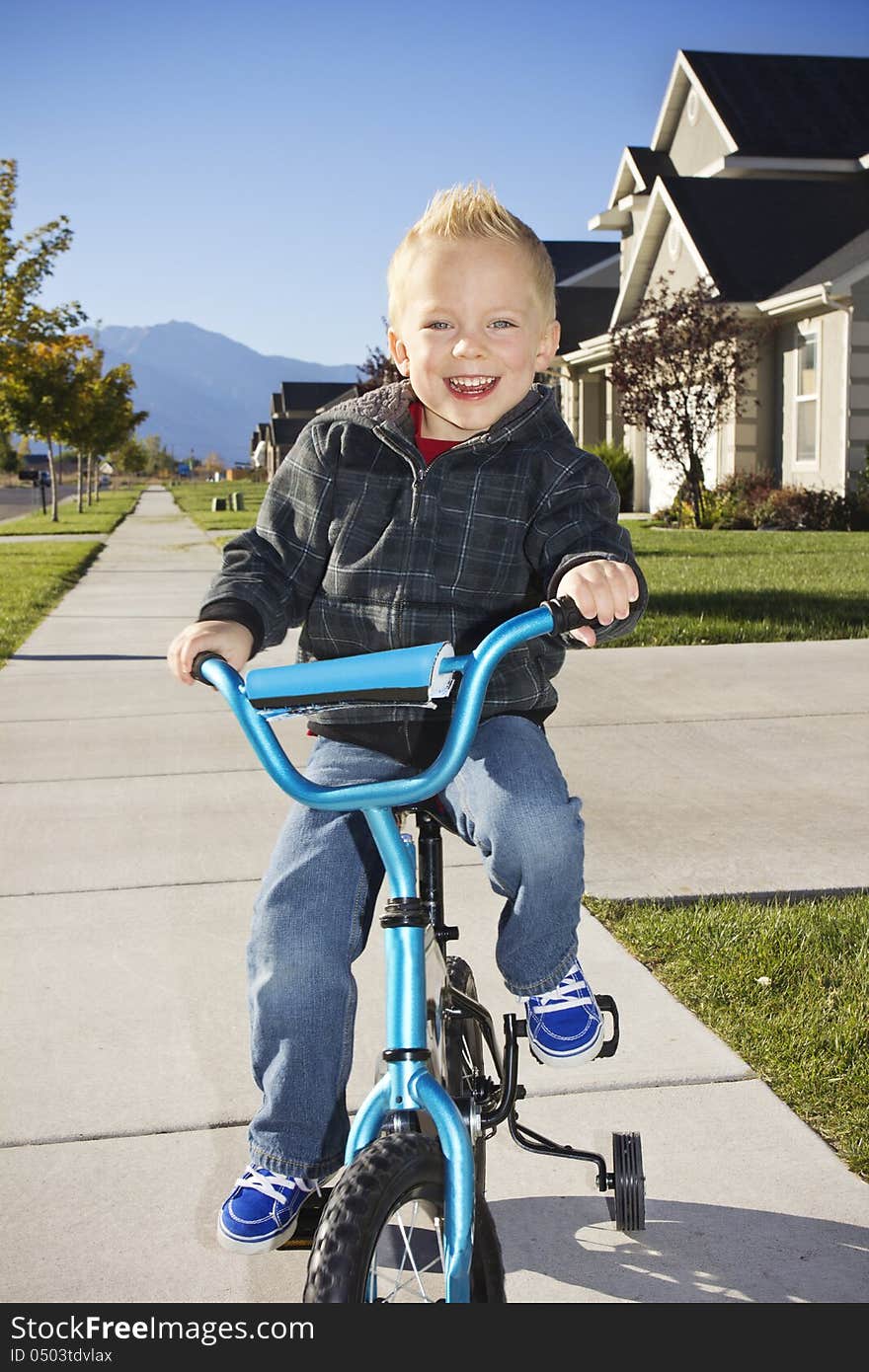 A cute Little boy learning to ride a bike with training wheels on the sidewalk in front of his house