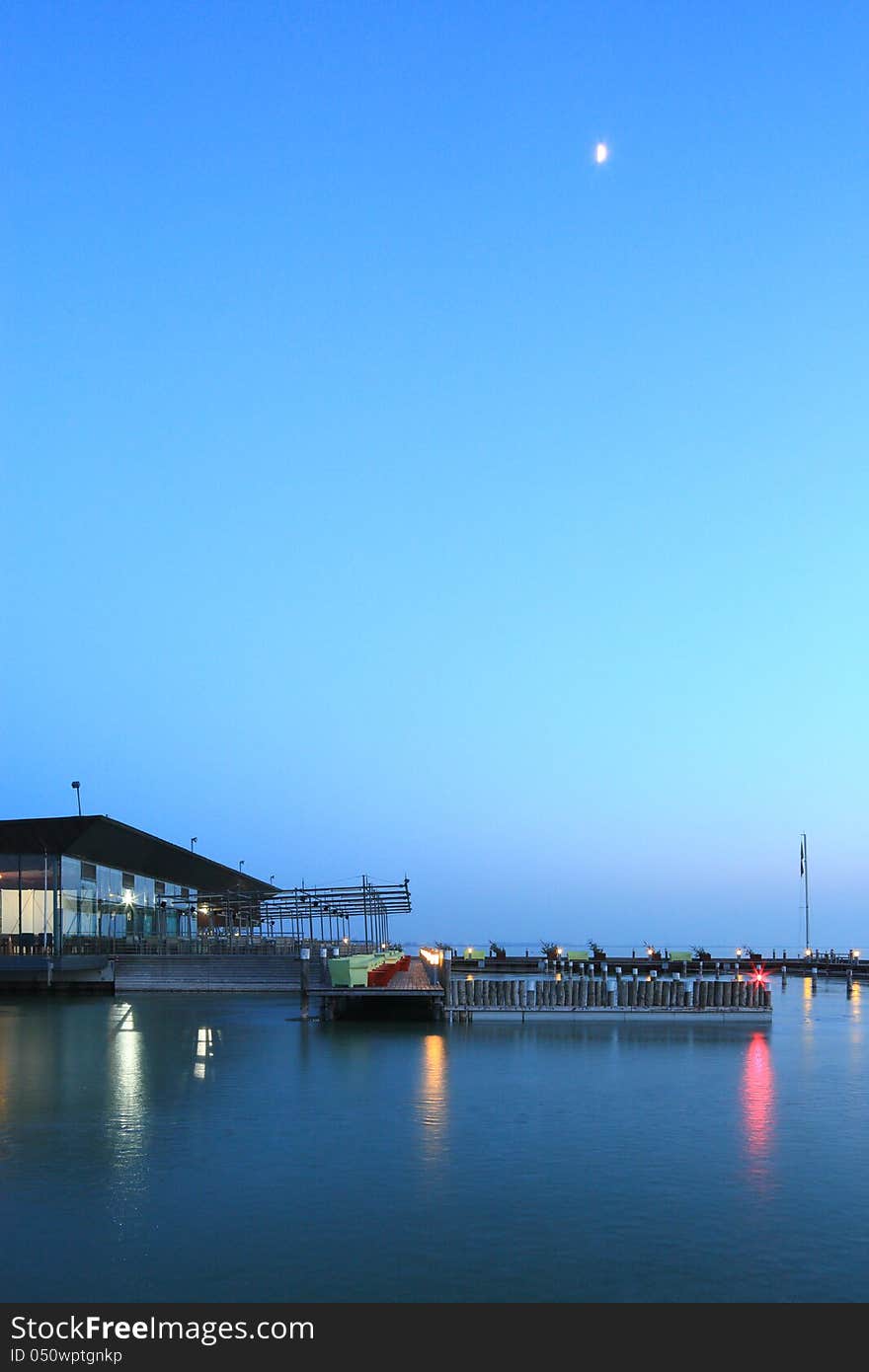 Landing stage on a big lake. The water is frozen, the stage deserted, also the bar on the lefthand. The sky is blue after sunset. The moon has risen high up. Landing stage on a big lake. The water is frozen, the stage deserted, also the bar on the lefthand. The sky is blue after sunset. The moon has risen high up.