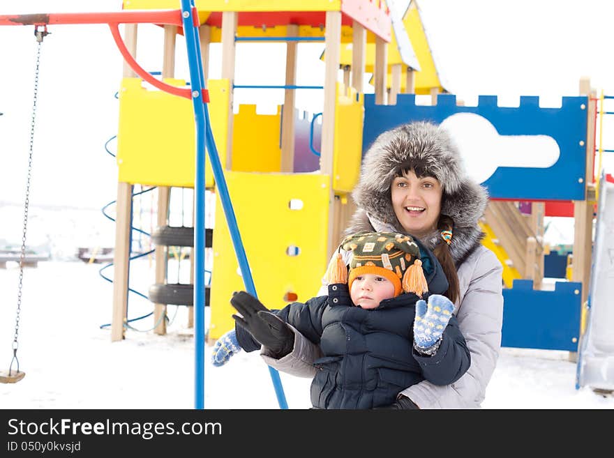 Happy mother and her young son standing in winter outfits waving at the camera in front of colourful childrens playground equipment in the snow. Happy mother and her young son standing in winter outfits waving at the camera in front of colourful childrens playground equipment in the snow