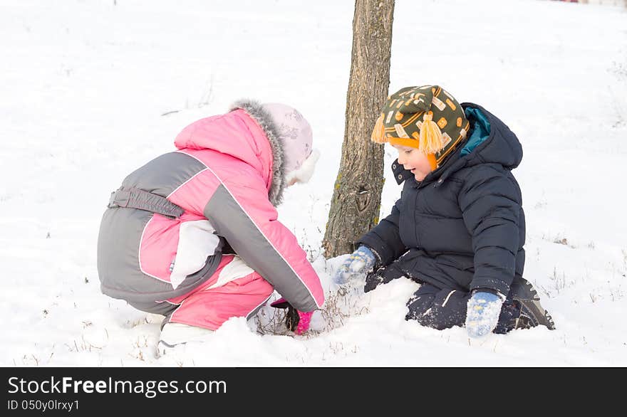 Two young children wrapped up warmly against the winter chill digging in the snow together at the foot of a small tree. Two young children wrapped up warmly against the winter chill digging in the snow together at the foot of a small tree