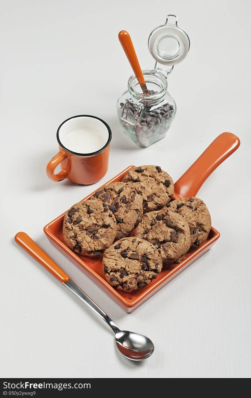 Top view of biscuits in orange ceramic pan, iar with chocolate drops, spoons and cup of milk on white wooden background. Top view of biscuits in orange ceramic pan, iar with chocolate drops, spoons and cup of milk on white wooden background