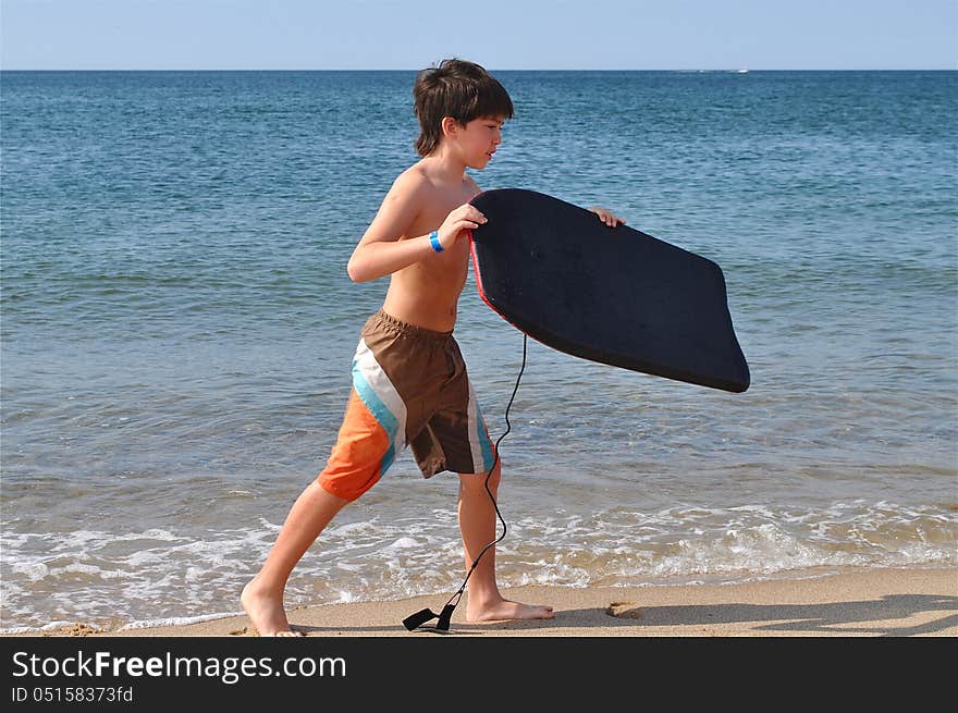 A young boy is walking in the shore at a beach with a board, ready to have great time in the water. A young boy is walking in the shore at a beach with a board, ready to have great time in the water.