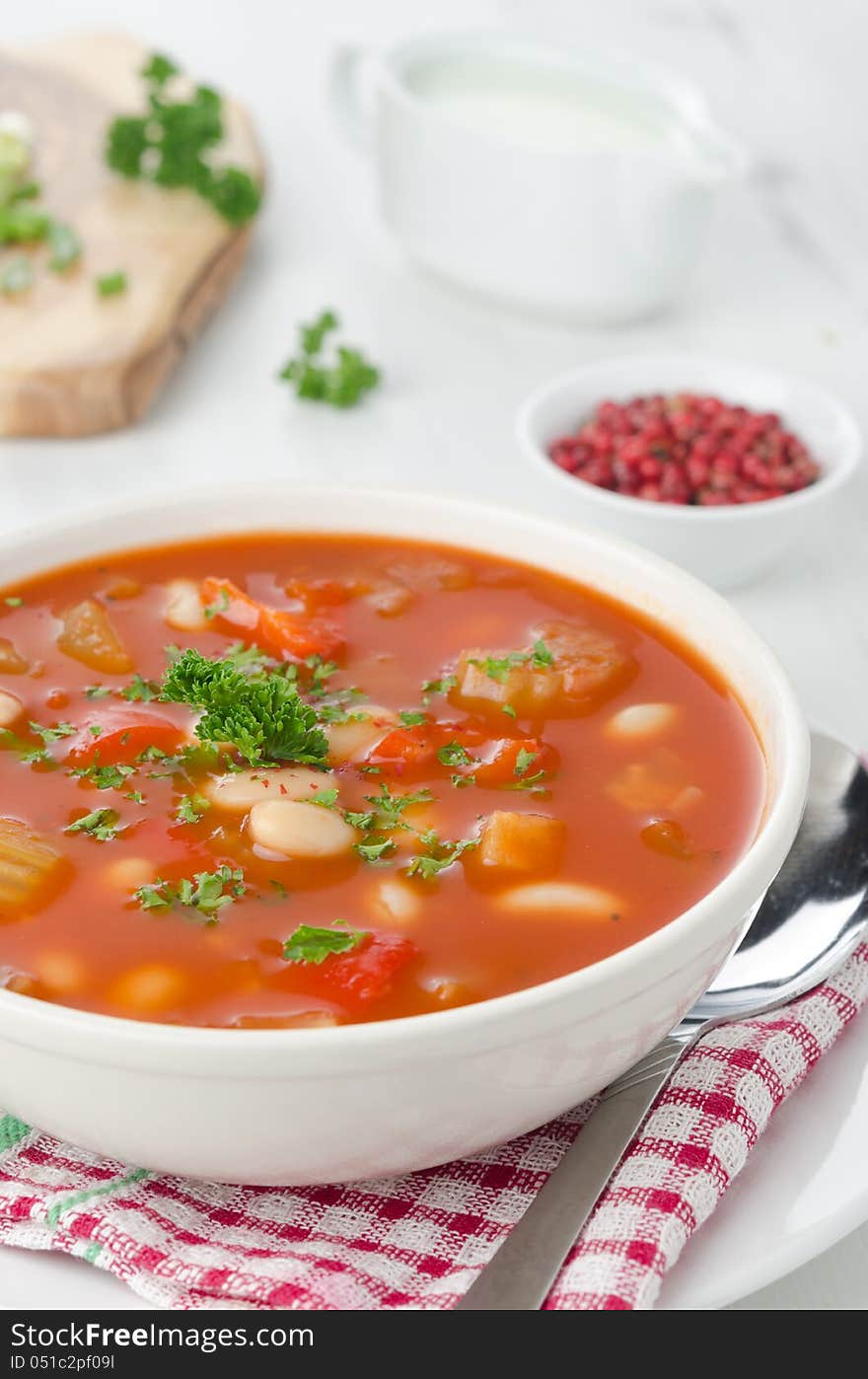 Bowl of roasted tomato soup with beans, celery and bell pepper, pink pepper and sour cream in the background