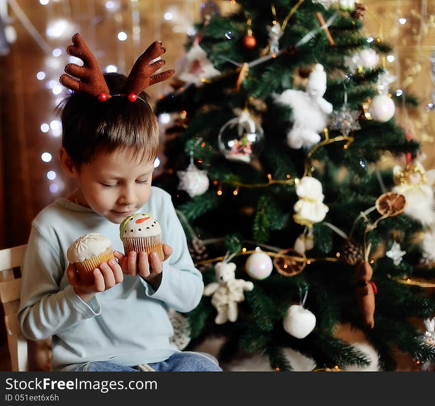 The boy with small horns of a deer under a New Year tree with beautiful fruitcakes in hand. The boy with small horns of a deer under a New Year tree with beautiful fruitcakes in hand