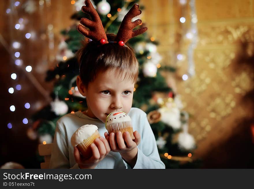 The boy with small horns of a deer under a New Year tree with beautiful fruitcakes in hand. The boy with small horns of a deer under a New Year tree with beautiful fruitcakes in hand