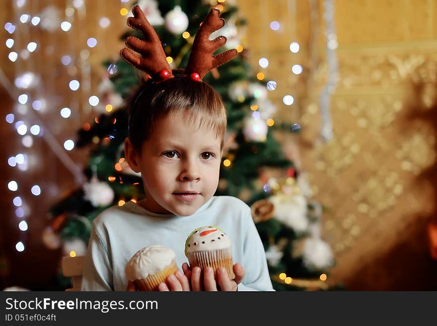 The boy with small horns of a deer under a New Year tree with beautiful fruitcakes in hand. The boy with small horns of a deer under a New Year tree with beautiful fruitcakes in hand