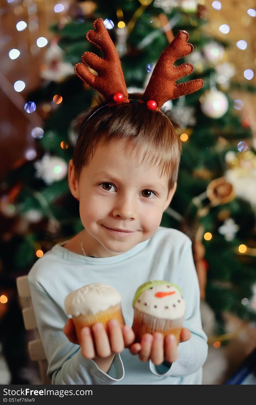 The boy with small horns of a deer under a New Year tree with beautiful fruitcakes in hand. The boy with small horns of a deer under a New Year tree with beautiful fruitcakes in hand