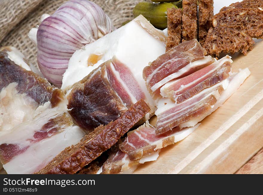 Raw Smoked Homemade Bacon Full Body and Slices with Garlic, Brown Bread and Gherkins closeup on Cutting Board. Raw Smoked Homemade Bacon Full Body and Slices with Garlic, Brown Bread and Gherkins closeup on Cutting Board