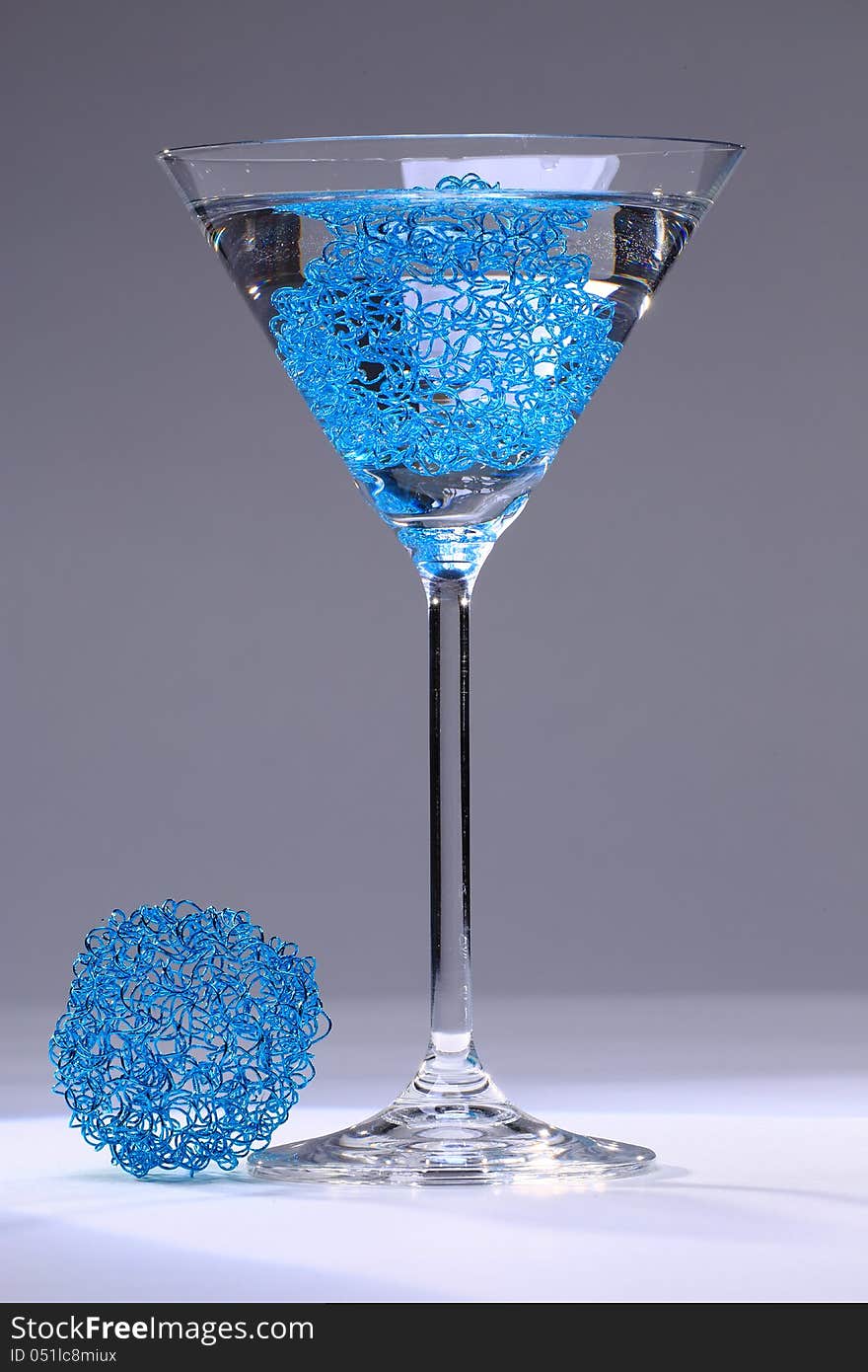 Cocktail glass formed like a cone on a stem filled with transparent fluid and a light blue mesh, in front of neutral grey background, a ball of blue mesh lying beneath it. Cocktail glass formed like a cone on a stem filled with transparent fluid and a light blue mesh, in front of neutral grey background, a ball of blue mesh lying beneath it