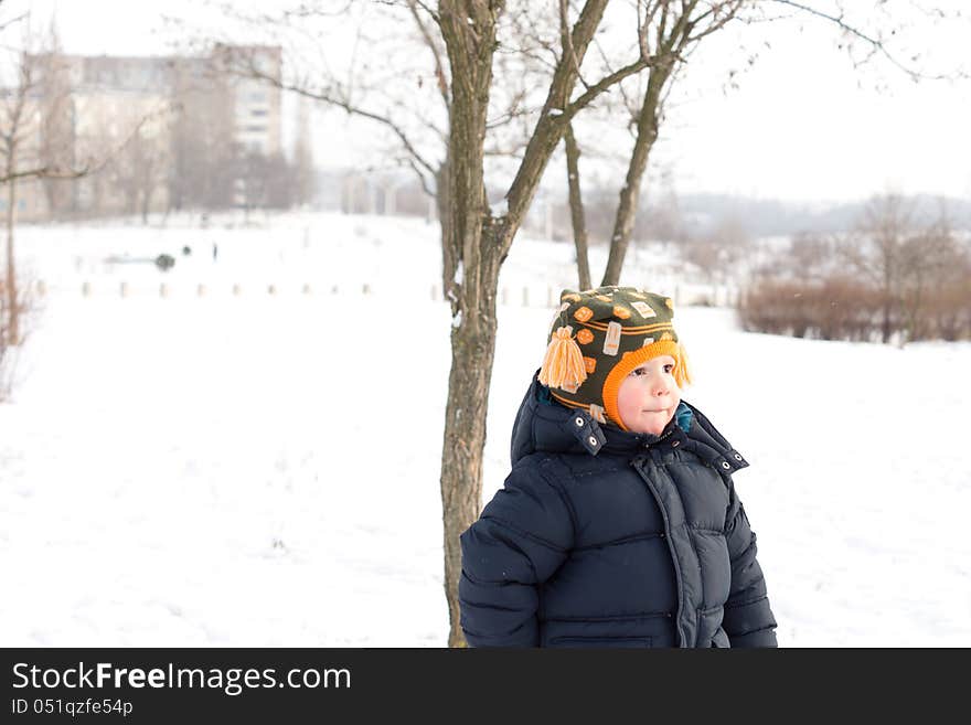 Small boy outdoors in winter snow standing alone against a small tree watching and waiting for someone to play with