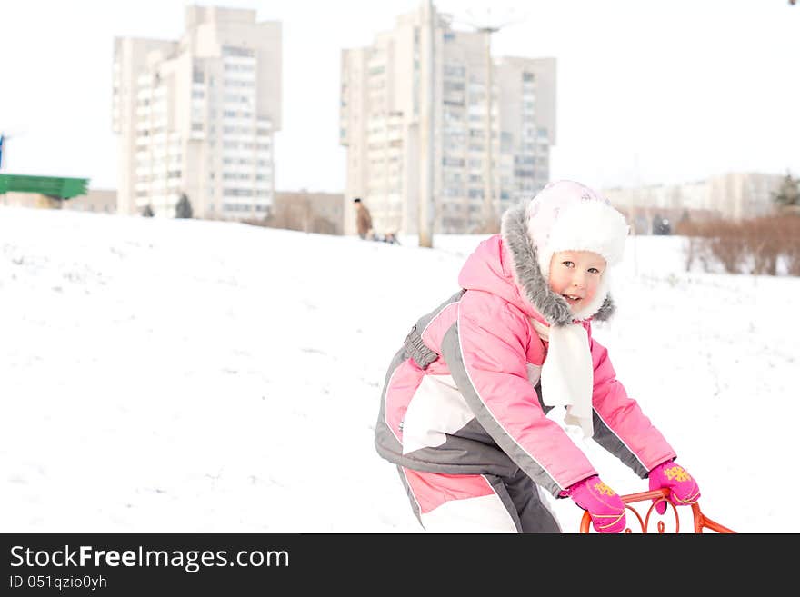 Pretty little girl in a cute furry pink outfit playing with a sled in a snowy field with two apartment blocks visible in the distance on the horizon. Pretty little girl in a cute furry pink outfit playing with a sled in a snowy field with two apartment blocks visible in the distance on the horizon.