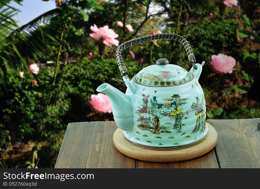 Chinese teapot standing in the garden of roses is on a wooden table on a round pedestal. Chinese teapot standing in the garden of roses is on a wooden table on a round pedestal