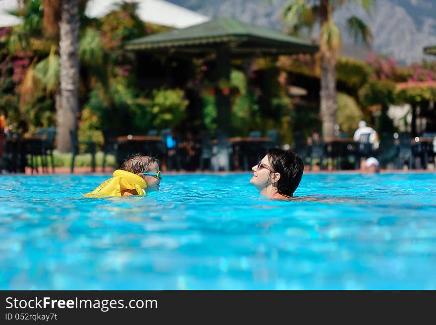 In the sunny day in the summer the boy in sun glasses and in a life jacket plays pool with mum. In the sunny day in the summer the boy in sun glasses and in a life jacket plays pool with mum