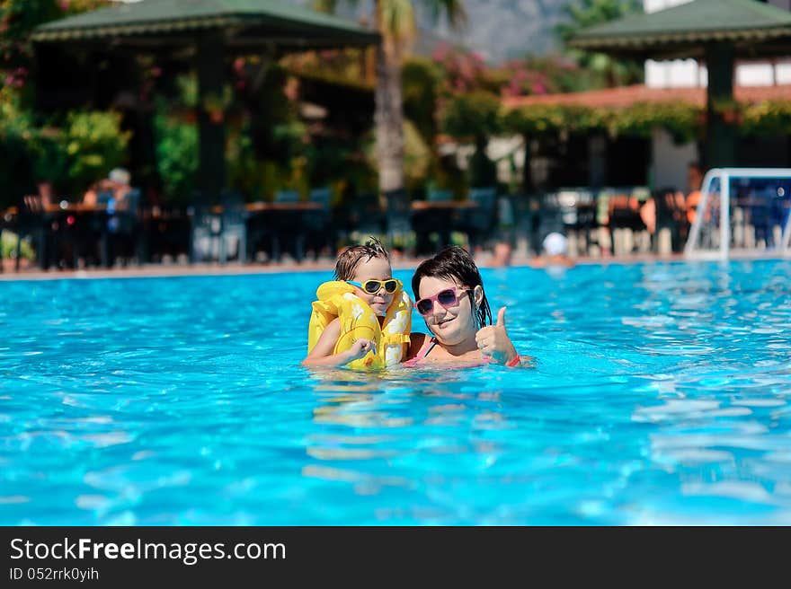 In the sunny day in the summer the boy in sun glasses and in a life jacket plays pool with mum. In the sunny day in the summer the boy in sun glasses and in a life jacket plays pool with mum