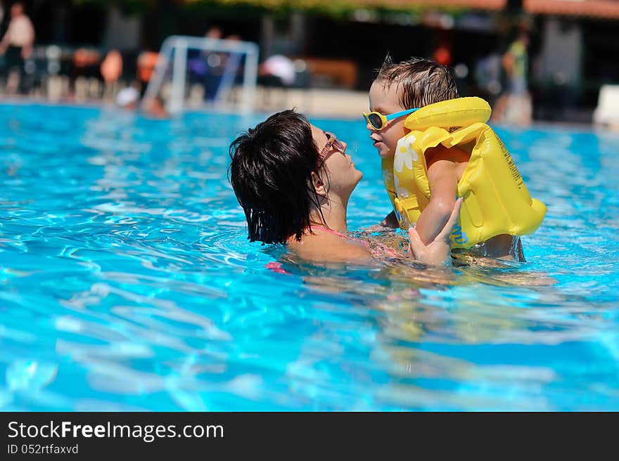 In the sunny day in the summer the boy in sun glasses and in a life jacket plays pool with mum. In the sunny day in the summer the boy in sun glasses and in a life jacket plays pool with mum