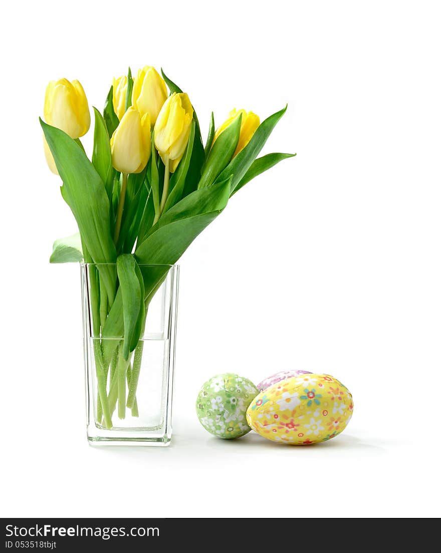 Studio macro of yellow tulips (Tulipa gesneriana) in a glass vase with decorative paper covered easter eggs against a white background. Copy space. Studio macro of yellow tulips (Tulipa gesneriana) in a glass vase with decorative paper covered easter eggs against a white background. Copy space.