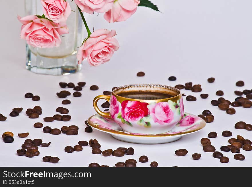 Small cup of coffee from an old service that scattered coffee beans and the background - a vase of beautiful roses. Small cup of coffee from an old service that scattered coffee beans and the background - a vase of beautiful roses