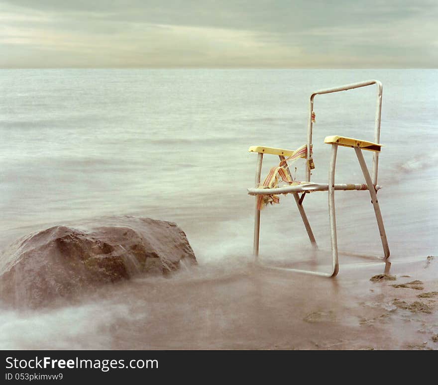 Old wrecked empty frame lawn chair sitting on the shore next to a boulder, with a cloudy sky. Old wrecked empty frame lawn chair sitting on the shore next to a boulder, with a cloudy sky.