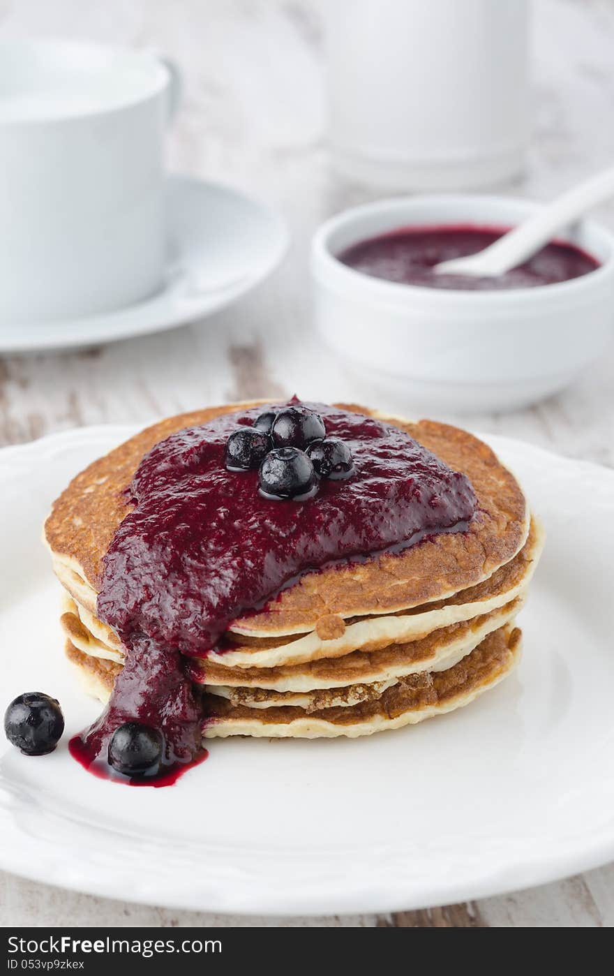 Stack of pancakes with black currant jam on a white plate, a bowl of jam in the background. Stack of pancakes with black currant jam on a white plate, a bowl of jam in the background