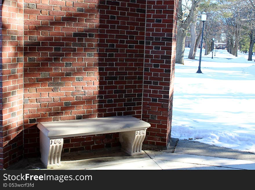 Stone seat in sunshine and curve of red brick wall with view of snowy walkway,line of trees and lampost. Stone seat in sunshine and curve of red brick wall with view of snowy walkway,line of trees and lampost.