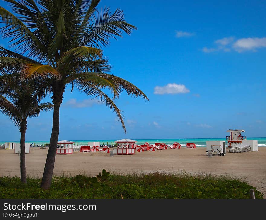 A wide stretch of beach along the vibrant, turquoise blue waters of the atlantic ocean with palm trees and colorful red and white beach umbrellas, lounges, and huts. A wide stretch of beach along the vibrant, turquoise blue waters of the atlantic ocean with palm trees and colorful red and white beach umbrellas, lounges, and huts.