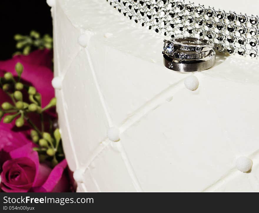 Wedding rings atop a bejeweled white wedding cake with pink roses as a background. Wedding rings atop a bejeweled white wedding cake with pink roses as a background.