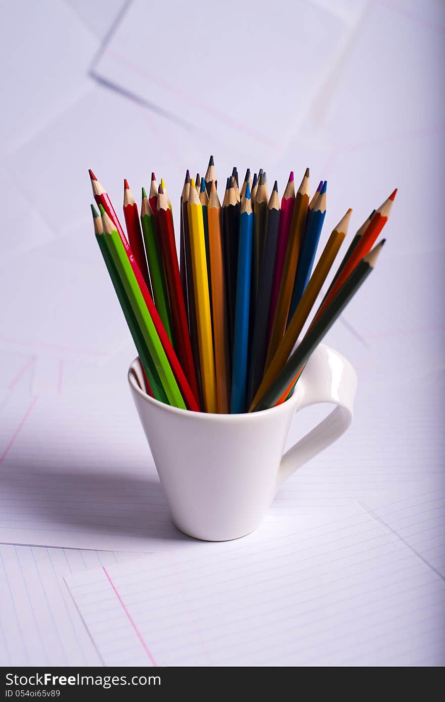 Many color pencils standing in a white mug against from writing-book sheets