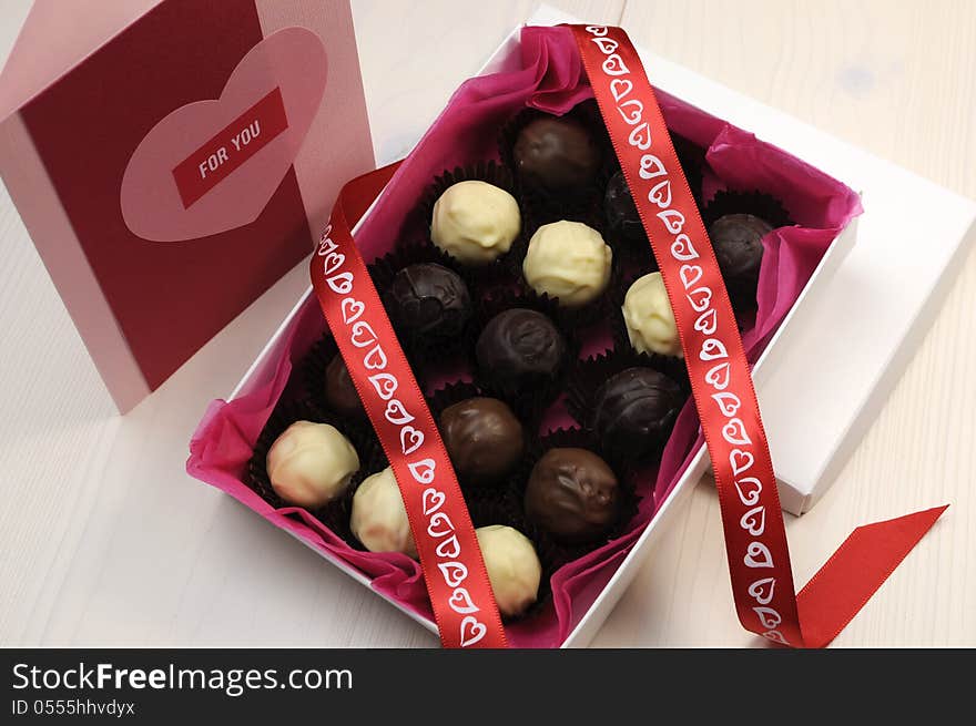 Box of chocolates with red heart ribbon and pink and red handmade gift card on white natural wood table. Box of chocolates with red heart ribbon and pink and red handmade gift card on white natural wood table.