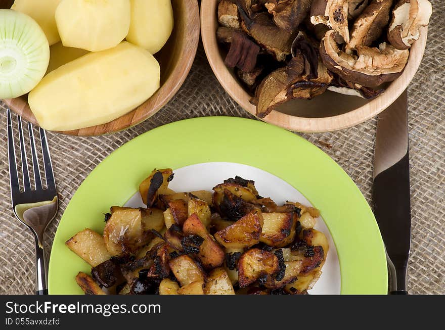 Traditional Roasted Potato with Mushrooms, Onion and Spices. Arrangement of Green Plate with Prepared Potato, Two Wood Bowls with Raw Potato, Onion and Slices of Mushrooms and Fork with Knife close up