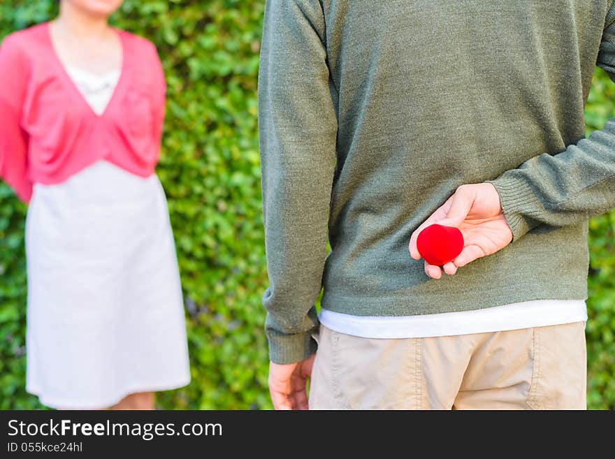 Man hide red heart shape wedding ring box on his back with a women in background. Man hide red heart shape wedding ring box on his back with a women in background