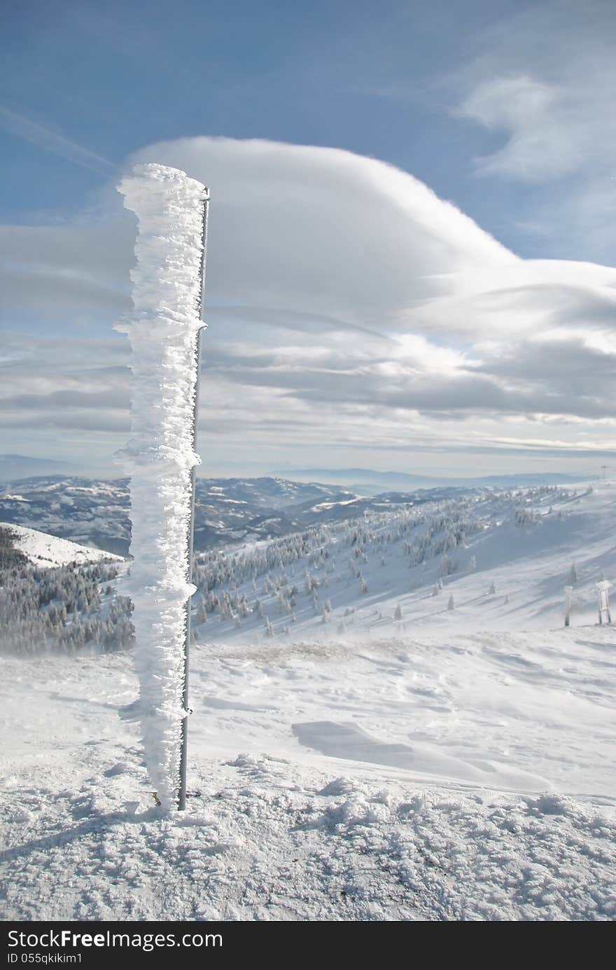 Top of the mountain in winter with iron pillar covered by icicles and white clouds on blue sky. Top of the mountain in winter with iron pillar covered by icicles and white clouds on blue sky